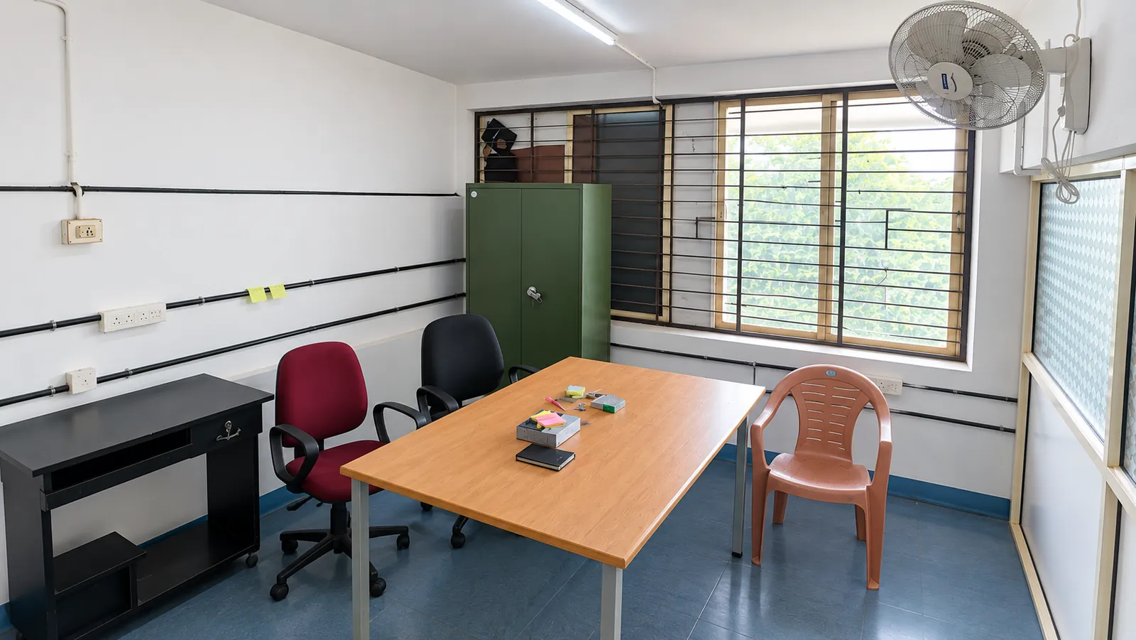 Cabin 2 at TBI MESCE: a wooden table with sticky notes, red and black office chairs, a plastic chair, a computer desk and a green steel cupboard beside a large window