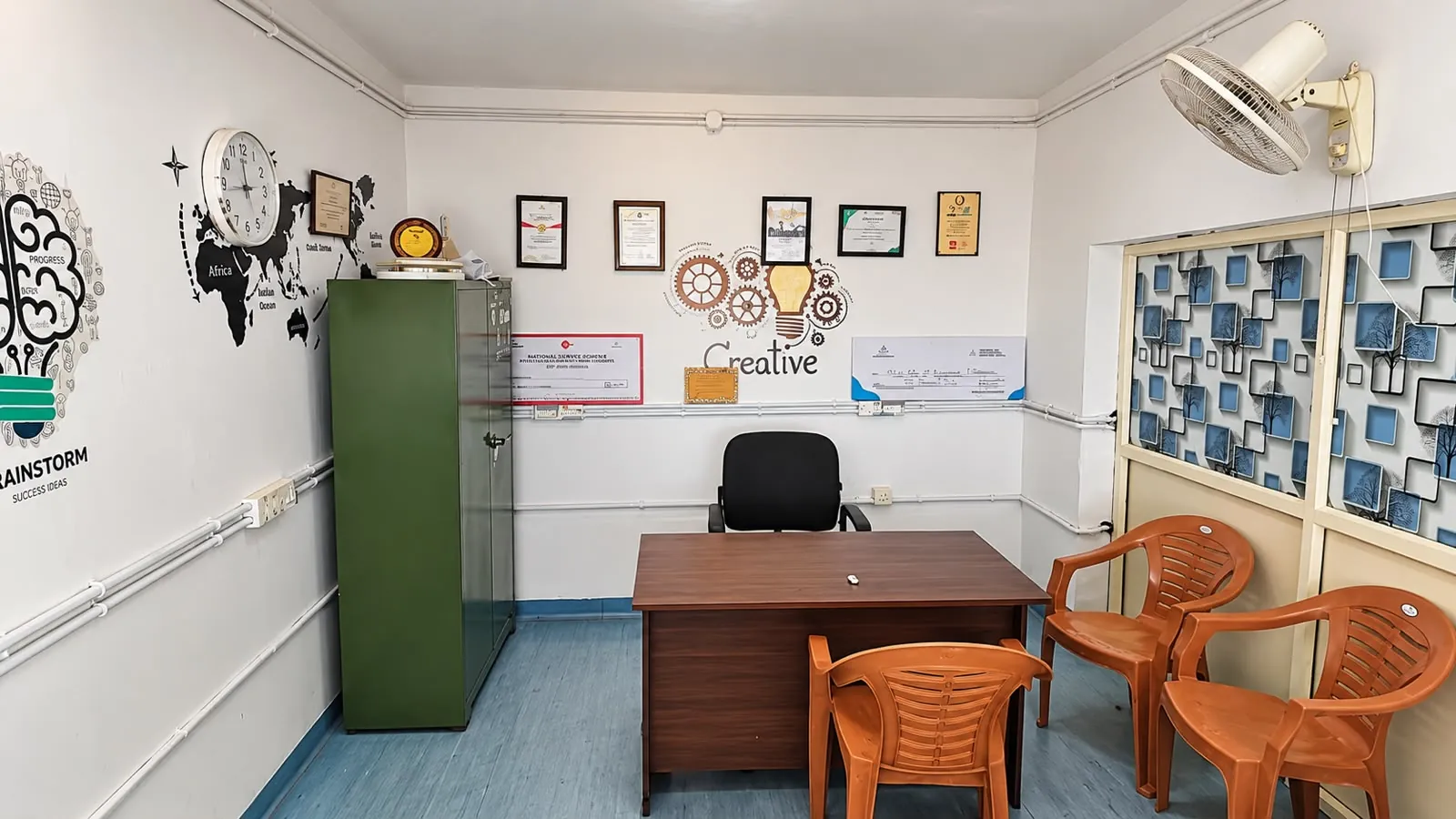 A room at TBI MESCE with a desk and office chair, a green steel cupboard, orange visitor chairs, framed certificates and awards, and a "Creative" gears-and-lightbulb mural on the wall